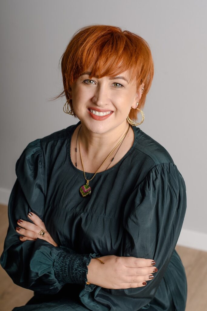 Julia Piotrowska-Haładuda, psycholożka i psychotraumatolożka, siedzi z założonymi rękami i uśmiecha się.